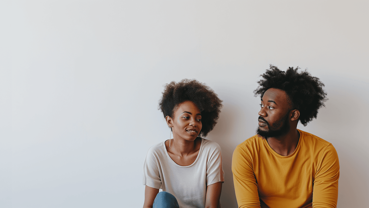 A couple sitting side by side against a white wall, glancing at each other thoughtfully